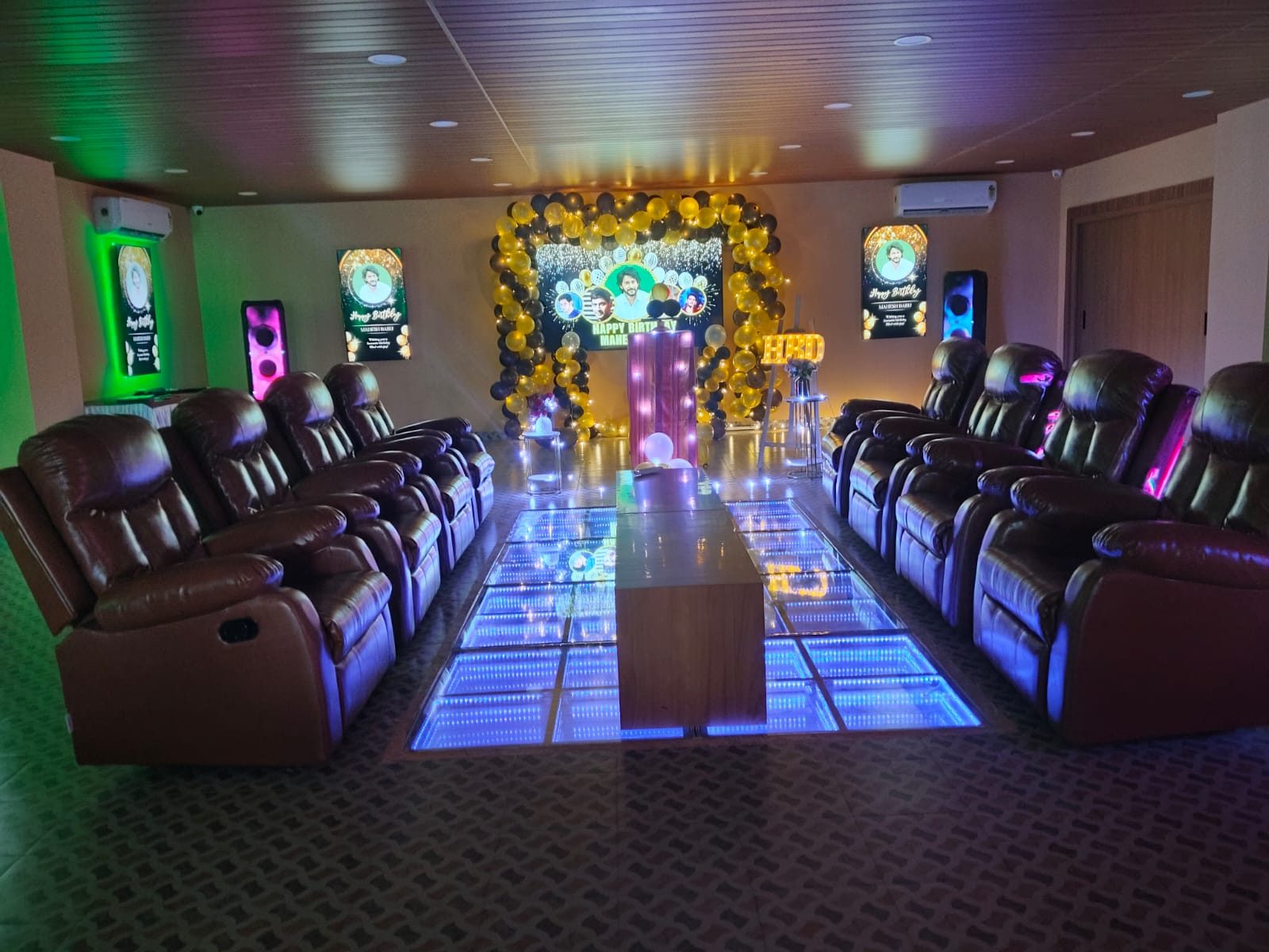 Party hall set for a birthday — recliners along a lit LED dance floor, balloon arch and glowing HBD letters